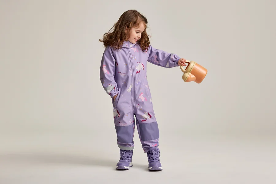 Mädchen mit braunen, lockigen Haaren in lila Overall mit Einhornmuster und lila Schuhen hält eine orangefarbene Gießkanne.