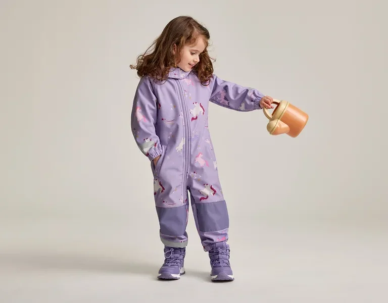 Mädchen mit braunen, lockigen Haaren in lila Overall mit Einhornmuster und lila Schuhen hält eine orangefarbene Gießkanne.