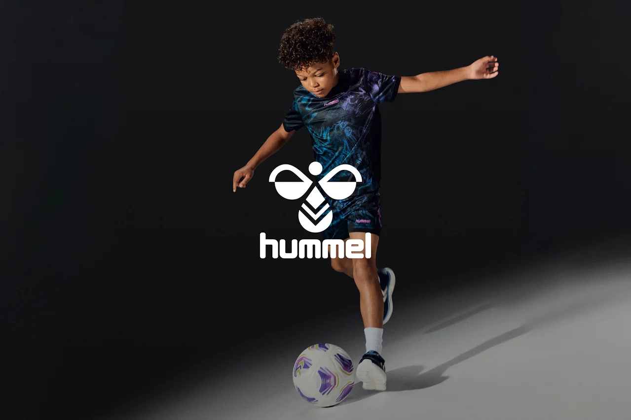Junger Junge in einem schwarz-lila gemusterten Sport-Outfit, der einen weiß-lila Fußball tritt. Ein weißes Hummel-Logo ist überlagert.