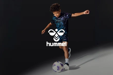 Junger Junge in dunklem, gemustertem T-Shirt und Shorts, der einen weiß-violetten Fußball kickt, mit dem weißen Hummel-Logo.