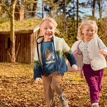 Zwei lächelnde Kinder, ein Junge und ein Mädchen, laufen durch eine laubbedeckte Naturlandschaft. Der Junge trägt ein blaues Shirt mit Motiv, eine farbige Fleecejacke und braune Cordhosen. Das Mädchen trägt ein gemustertes Shirt, eine cremefarbene flauschige Weste und magentafarbene Cordhosen. Im Hintergrund ist ein Holzhaus mit Grasdach zu sehen.