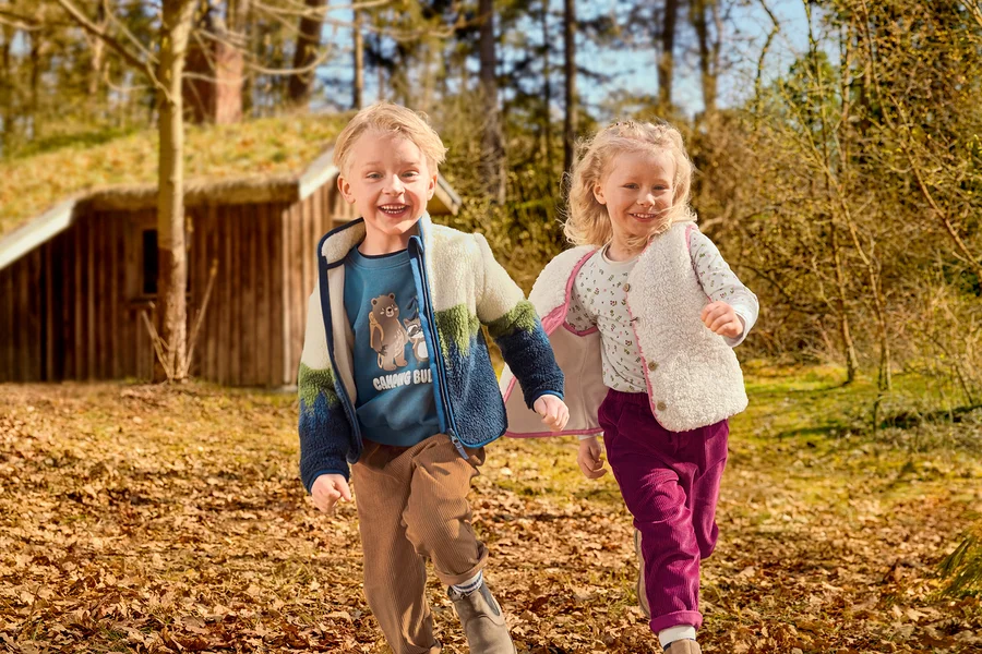 Zwei lächelnde Kinder, ein Junge und ein Mädchen, laufen durch eine laubbedeckte Naturlandschaft. Der Junge trägt ein blaues Shirt mit Motiv, eine farbige Fleecejacke und braune Cordhosen. Das Mädchen trägt ein gemustertes Shirt, eine cremefarbene flauschige Weste und magentafarbene Cordhosen. Im Hintergrund ist ein Holzhaus mit Grasdach zu sehen.
