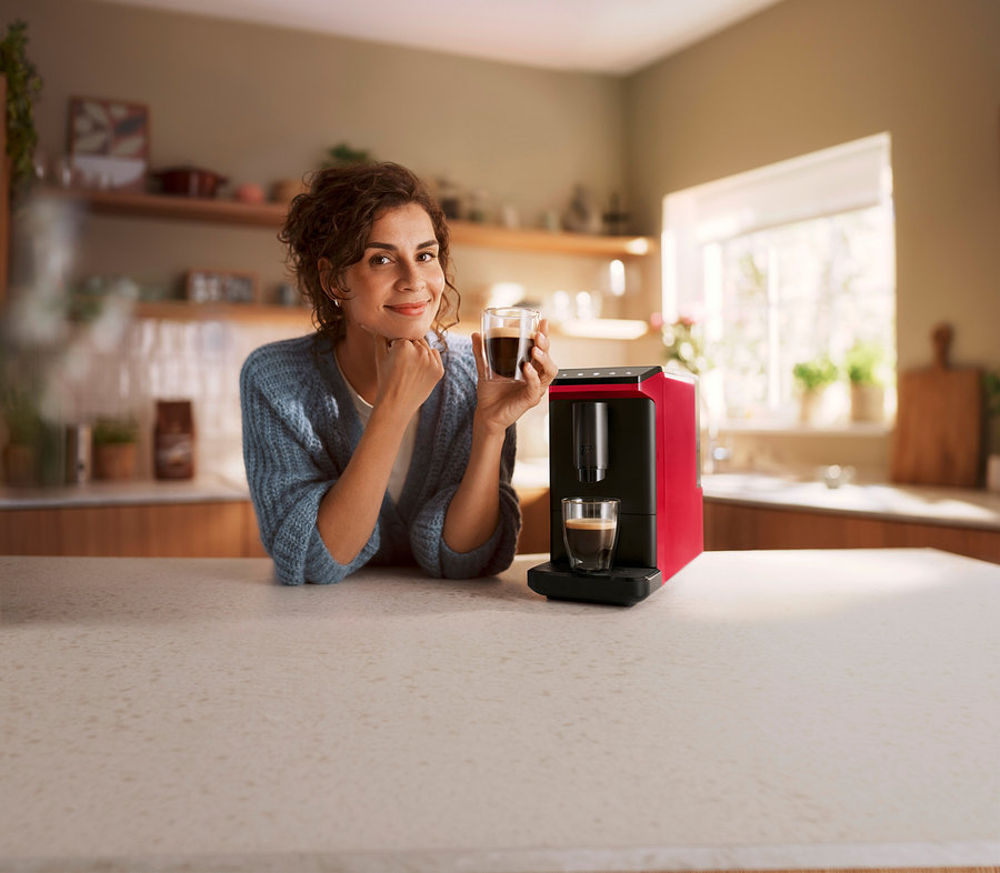 Lächelnde Frau lehnt an einer Küchenzeile und hält eine Tasse Kaffee, neben ihr ein roter Kaffeevollautomat »Esperto mini«, Lovely Red, der eine weitere Tasse Kaffee zubereitet.