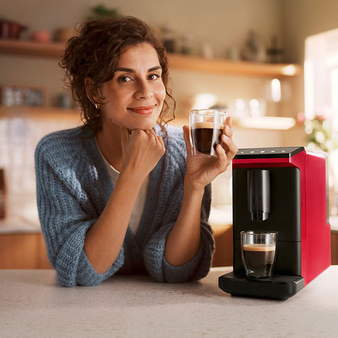 Lächelnde Frau lehnt an einer Küchenzeile und hält eine Tasse Kaffee, neben ihr ein roter Kaffeevollautomat »Esperto mini«, Lovely Red, der eine weitere Tasse Kaffee zubereitet.