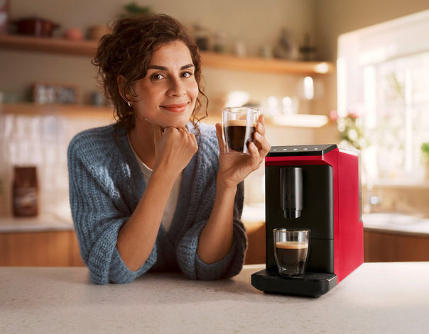 Lächelnde Frau lehnt an einer Küchenzeile und hält eine Tasse Kaffee, neben ihr ein roter Kaffeevollautomat »Esperto mini«, Lovely Red, der eine weitere Tasse Kaffee zubereitet.