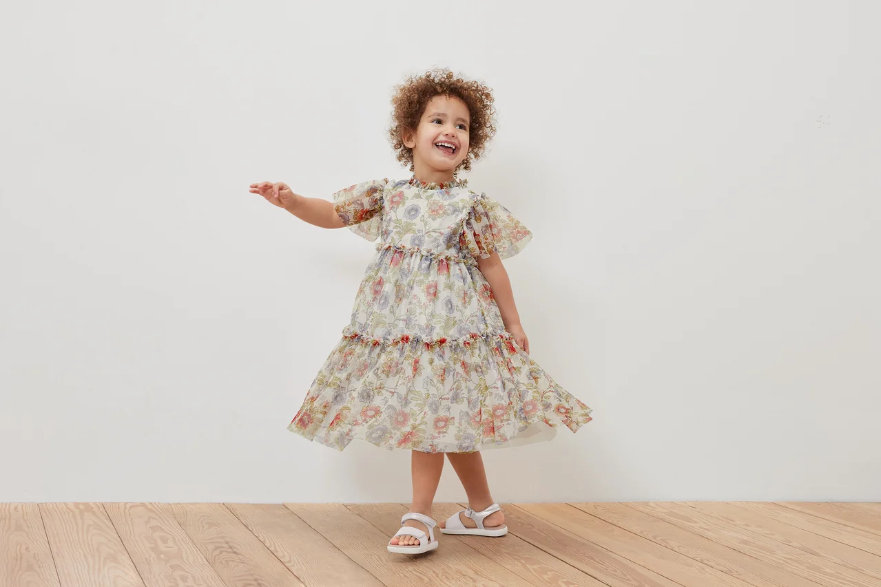 Ein lächelndes Mädchen posiert in einem geblümten Kinder-Tüllkleid und weißen Sandalen.