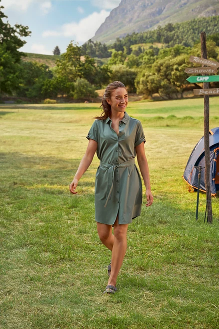 Frau geht auf einer Wiese in einem grünen Hemdkleid mit Gürtel. Im Hintergrund sind ein Zelt und ein Wegweiser zu sehen.