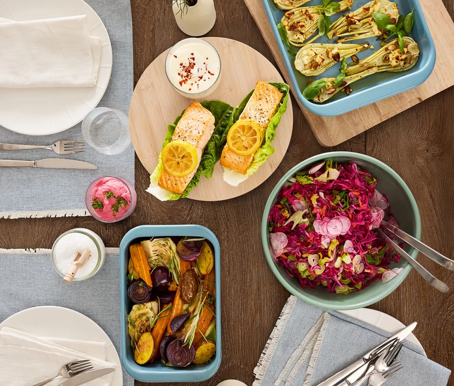 Draufsicht auf einen Holztisch mit verschiedenen Speisen, darunter Lachs, Gemüse und Salat.