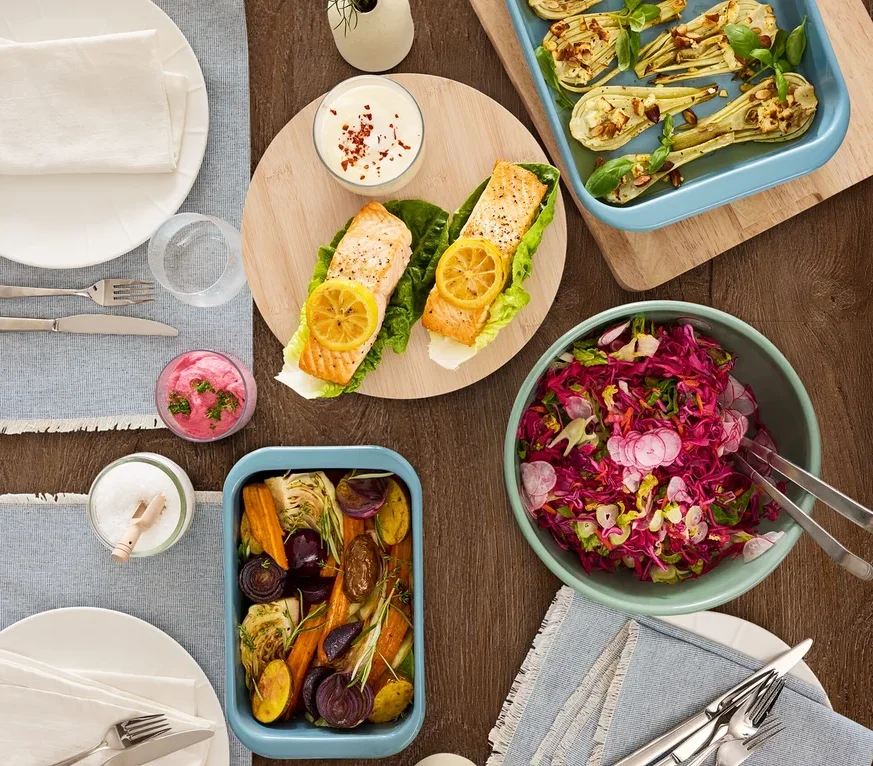 Draufsicht auf einen Holztisch mit verschiedenen Speisen, darunter Lachs, Gemüse und Salat.
