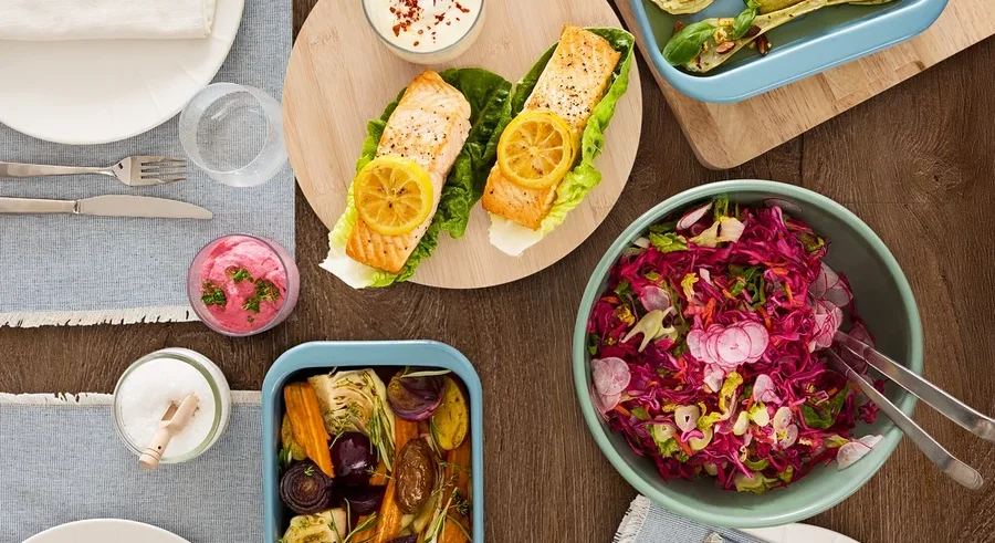 Draufsicht auf einen Holztisch mit verschiedenen Speisen, darunter Lachs, Gemüse und Salat.