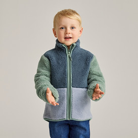 Ein blonder Junge mit blauen Augen trägt eine Fleecejacke in Blau, Grün und Hellblau und blaue Jeans.