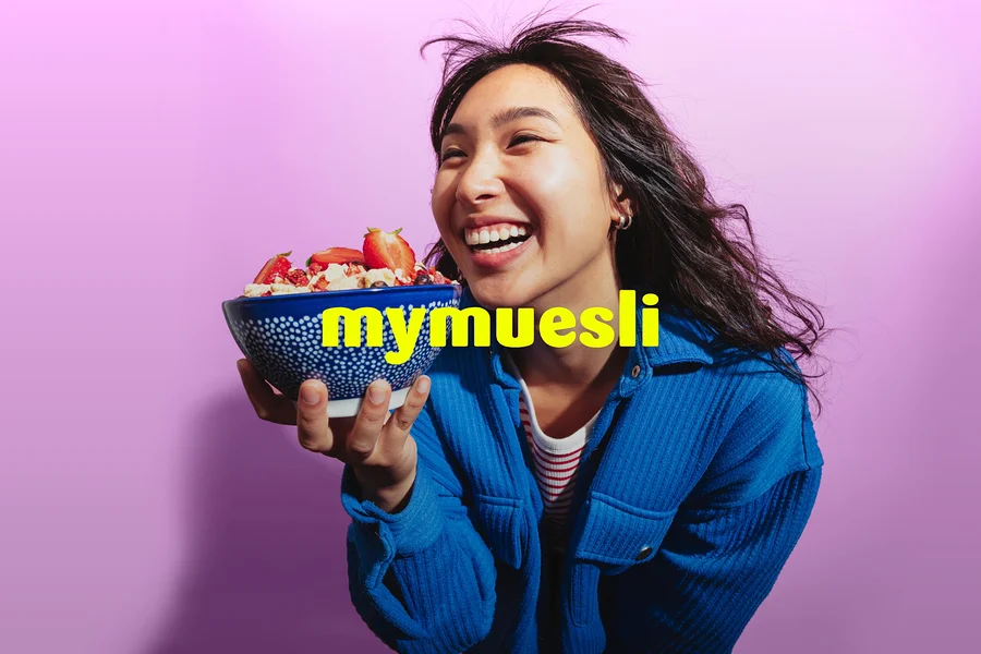 Eine lachende Frau in einer blauen Jacke hält eine blaue Schale Müsli mit Erdbeeren vor einem lila Hintergrund. Der gelbe Text 'mymuesli' ist überlagert.