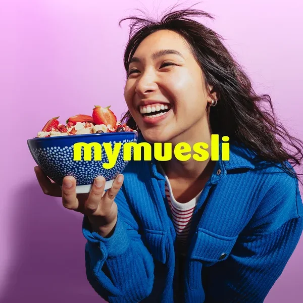 Eine lachende Frau in einer blauen Jacke hält eine blaue Schale Müsli mit Erdbeeren vor einem lila Hintergrund. Der gelbe Text 'mymuesli' ist überlagert.