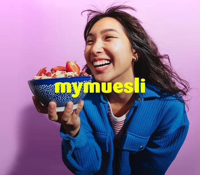 Eine lachende Frau in einer blauen Jacke hält eine blaue Schale Müsli mit Erdbeeren vor einem lila Hintergrund. Der gelbe Text 'mymuesli' ist überlagert.