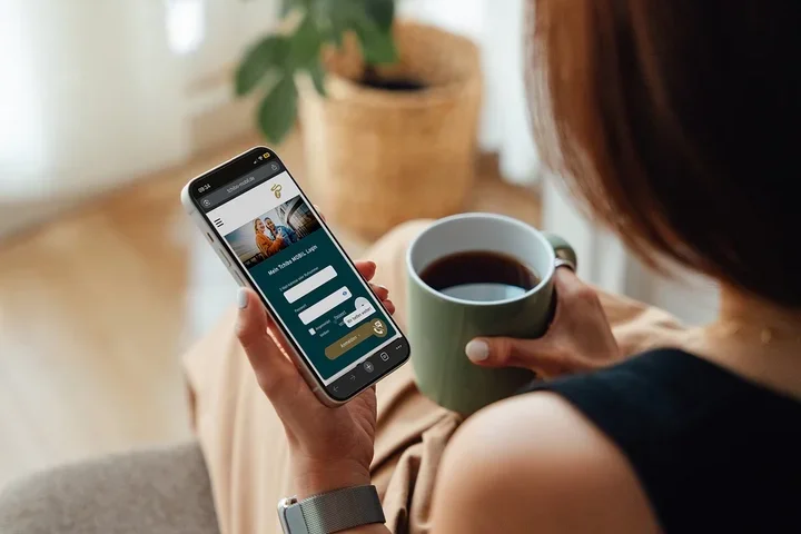 Eine Person von hinten hält ein Smartphone, das eine „Mein Tchibo MOBIL Login“-Seite anzeigt, und eine grüne Kaffeetasse. Sie trägt eine Armbanduhr.