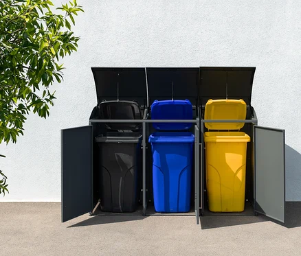 Drei farbige Mülltonnen in einer grauen Mülltonnenbox, 3er, stehen vor einer weißen Wand.