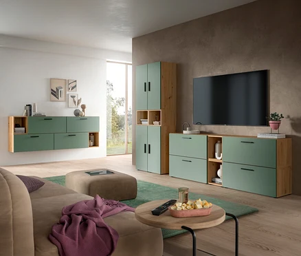 Modernes Wohnzimmer mit beiger Couch, rundem Couchtisch und großem Hocker. An der Wand sind schwebende Schränke und ein TV-Möbel in Salbeigrün und hellem Holz mit einem Fernseher.