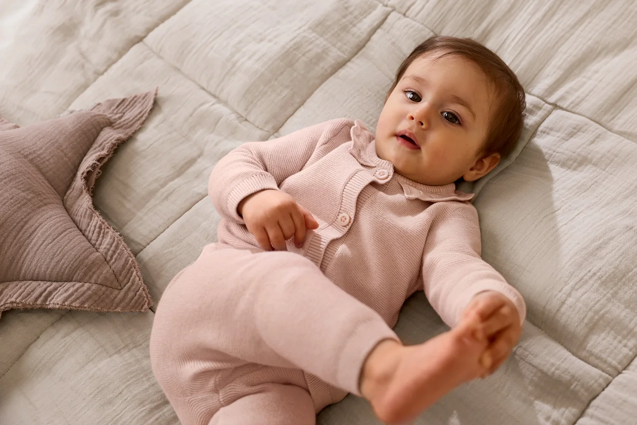Ein Baby in einem rosa Strick-Cardigan und passender Hose liegt auf einer hellgrauen, strukturierten Decke und blickt nach oben. Neben dem Baby liegt ein rosa sternförmiges Kissen.