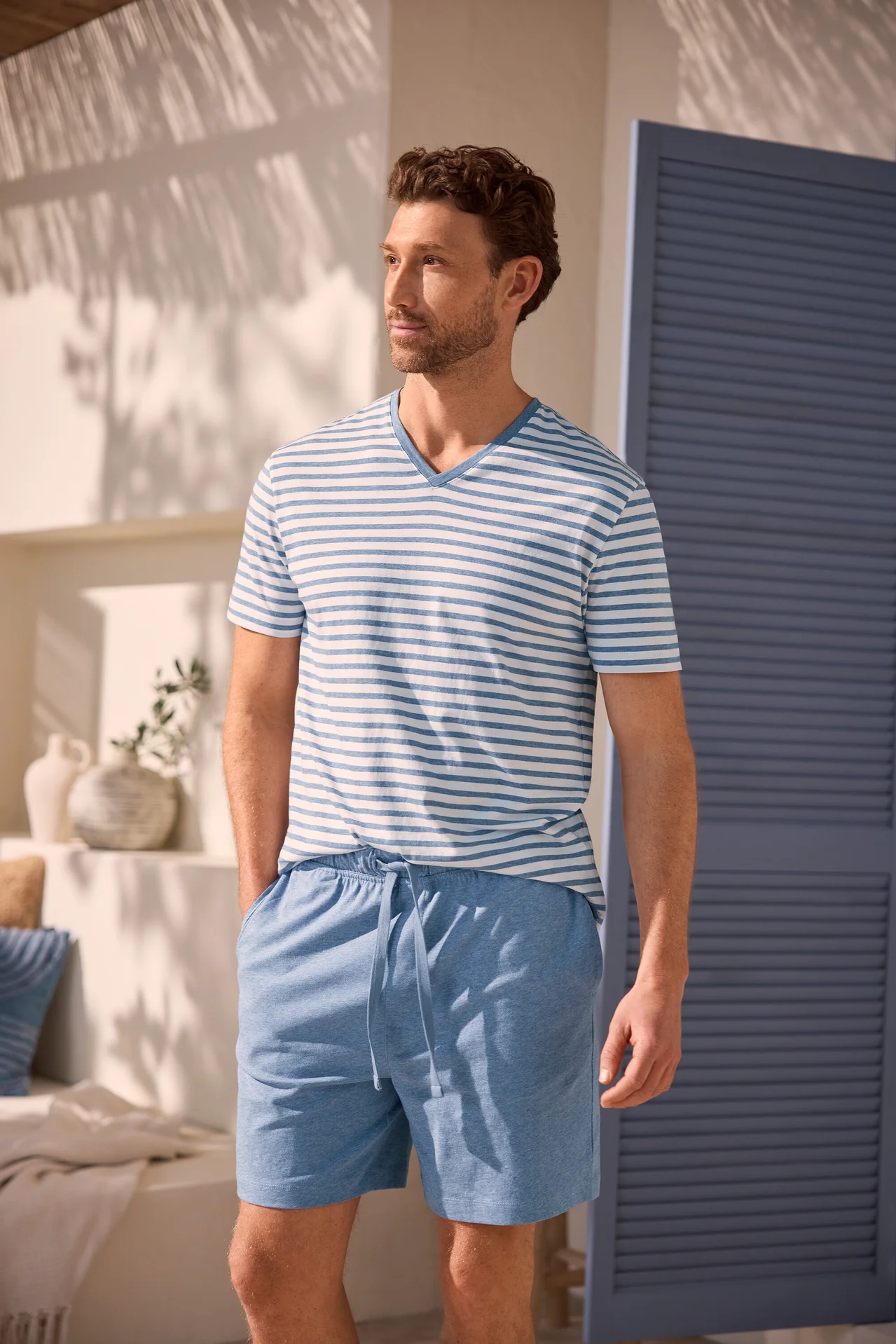 Ein Mann mit lockigem braunem Haar und Bart trägt ein blau-weiß gestreiftes V-Ausschnitt-T-Shirt und hellblaue Shorts. Er steht mit den Händen in den Taschen vor einer weißen Wand mit Schatten und einer blauen Tür.