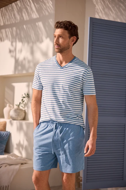 Ein Mann mit lockigem braunem Haar und Bart trägt ein blau-weiß gestreiftes V-Ausschnitt-T-Shirt und hellblaue Shorts. Er steht mit den Händen in den Taschen vor einer weißen Wand mit Schatten und einer blauen Tür.