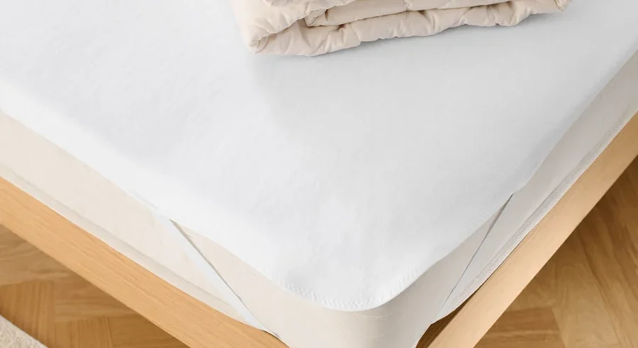 Auf einem Bett liegen ein weißer Matratzenschoner und eine gefaltete Decke.