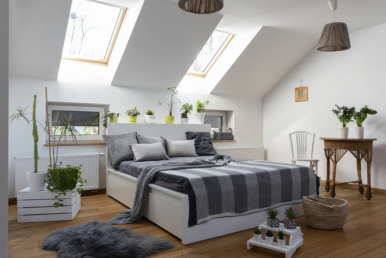 Schlafzimmer mit weißem Bett, grauen Kissen und Decke, sowie Pflanzen und Holzboden.