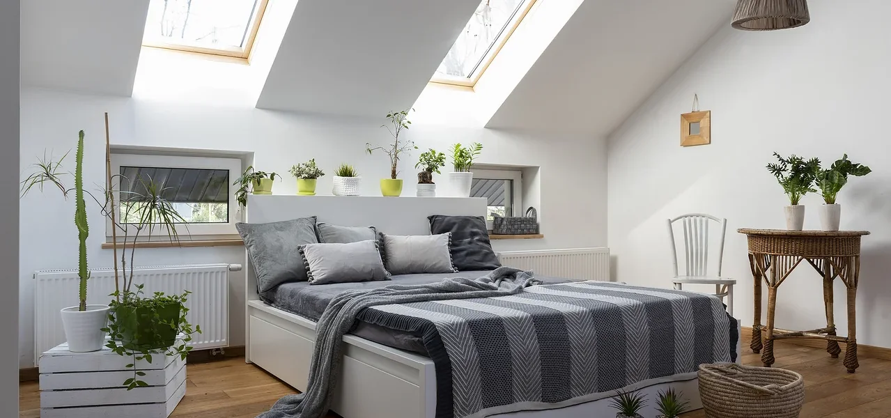 Schlafzimmer mit weißem Bett, grauen Kissen und Decke, sowie Pflanzen und Holzboden.