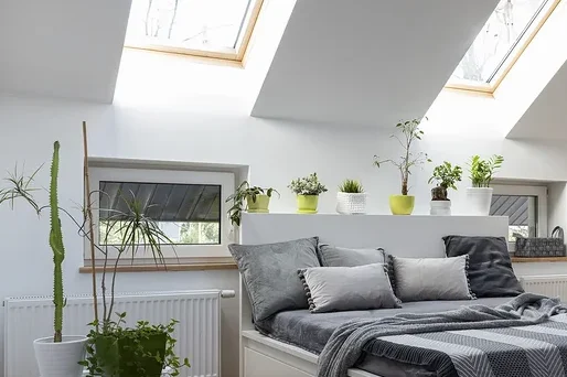 Schlafzimmer mit weißem Bett, grauen Kissen und Decke, sowie Pflanzen und Holzboden.