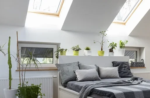 Schlafzimmer mit weißem Bett, grauen Kissen und Decke, sowie Pflanzen und Holzboden.