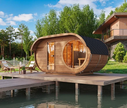 Hölzerne Fasssauna auf einer Holzterrasse über Wasser, mit Gartenmöbeln, umgeben von Bäumen und einem Haus im Hintergrund.