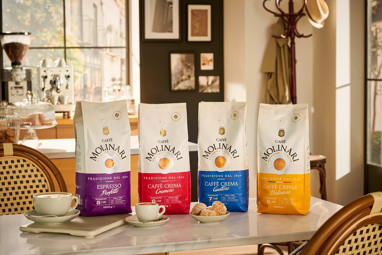 Vier Packungen Caffè Molinari stehen auf einem Tisch neben Tassen und Keksen.