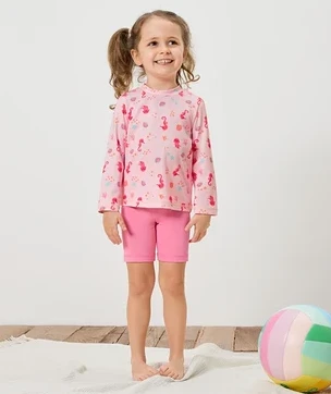 Ein Mädchen mit Zöpfen trägt ein rosa Langarmshirt mit Seepferdchenmuster und pinke Shorts. Ein bunter Ball liegt auf einem weißen Teppich.