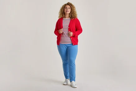 Eine Frau mit lockigem Haar steht und trägt einen offenen roten Cardigan über einem gestreiften T-Shirt, hellblauen Hosen und weißen Turnschuhen.