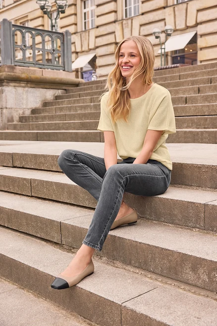 Eine lächelnde junge Frau mit blonden Haaren sitzt auf Steinstufen und trägt ein hellgelbes Oberteil, dunkelgraue Jeans und Leder-Ballerinas in Beige und Schwarz.