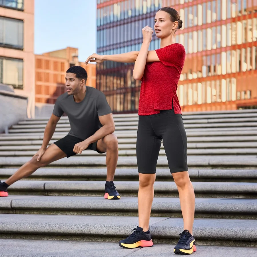 Frau in Sportradler und rotem Sportshirt dehnt sich auf Treppe, Mann in Sportshorts und grauem Funktionsshirt dehnt sich abseits.