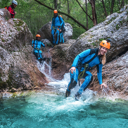 Fünf Personen beim Canyoning in einer felsigen Schlucht. Eine blonde Frau in blauem Neoprenanzug und orangefarbenem Helm lächelt, während sie ins türkise Wasser steigt. Dahinter rutscht eine Person einen kleinen Wasserfall hinunter, drei weitere stehen auf Felsen.