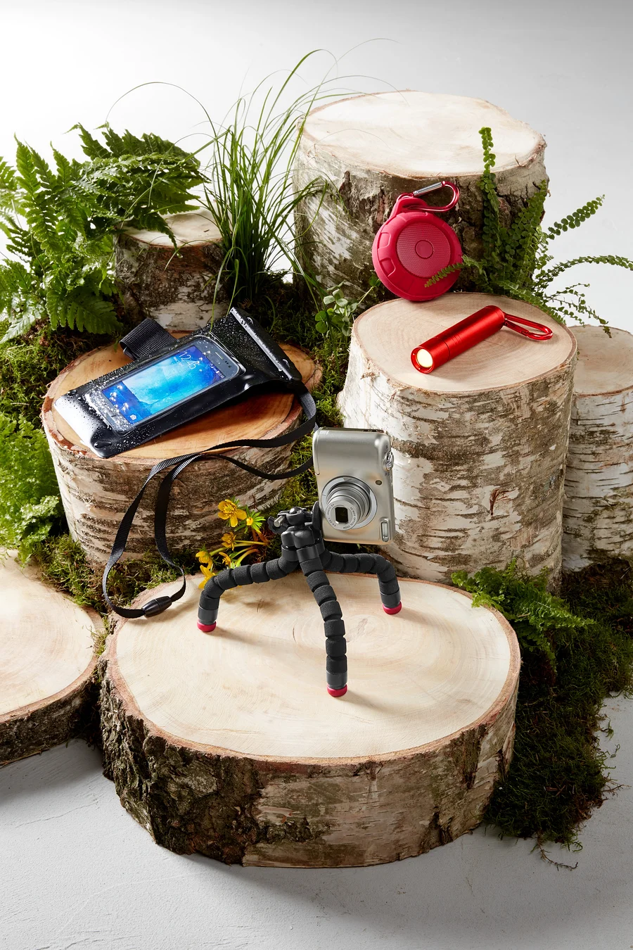 Kamera auf Stativ, wasserdichte Handyhülle, roter Lautsprecher und Taschenlampe auf Holzstümpfen mit Moos und Farnen.