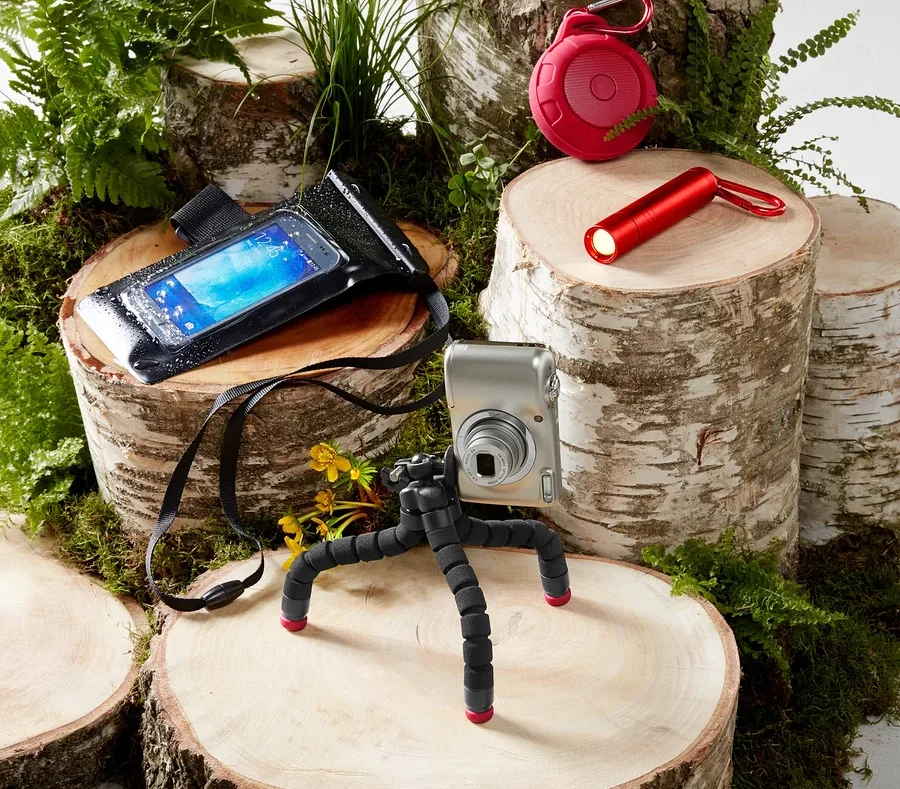 Kamera auf Stativ, wasserdichte Handyhülle, roter Lautsprecher und Taschenlampe auf Holzstümpfen mit Moos und Farnen.