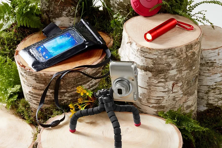 Kamera auf Stativ, wasserdichte Handyhülle, roter Lautsprecher und Taschenlampe auf Holzstümpfen mit Moos und Farnen.