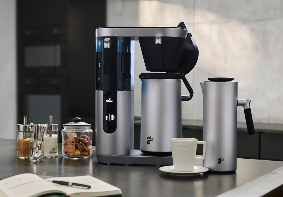 Silberne Profi-Filterkaffeemaschine Coffea Filbii neben Filbii 1L und 2L Thermoskannen. Auf dem Tisch liegen ein offenes Notizbuch und Gläser mit Zucker und Keksen.