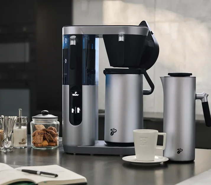Silberne Profi-Filterkaffeemaschine Coffea Filbii neben Filbii 1L und 2L Thermoskannen. Auf dem Tisch liegen ein offenes Notizbuch und Gläser mit Zucker und Keksen.