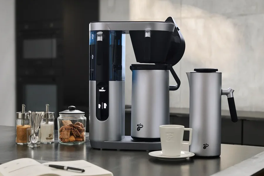 Silberne Profi-Filterkaffeemaschine Coffea Filbii neben Filbii 1L und 2L Thermoskannen. Auf dem Tisch liegen ein offenes Notizbuch und Gläser mit Zucker und Keksen.