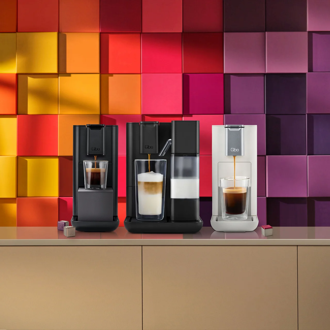 Drei Qbo Kaffeemaschinen in Schwarz und Weiss stehen auf einem Tresen vor einer bunten Wand, während Kaffee in Gläser gefüllt wird.
