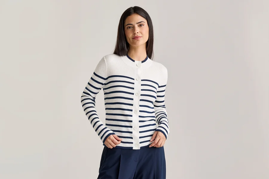 Frau mit dunkelbraunen Haaren trägt eine weiß-blau gestreifte Strickjacke mit Knöpfen und eine marineblaue Hose.