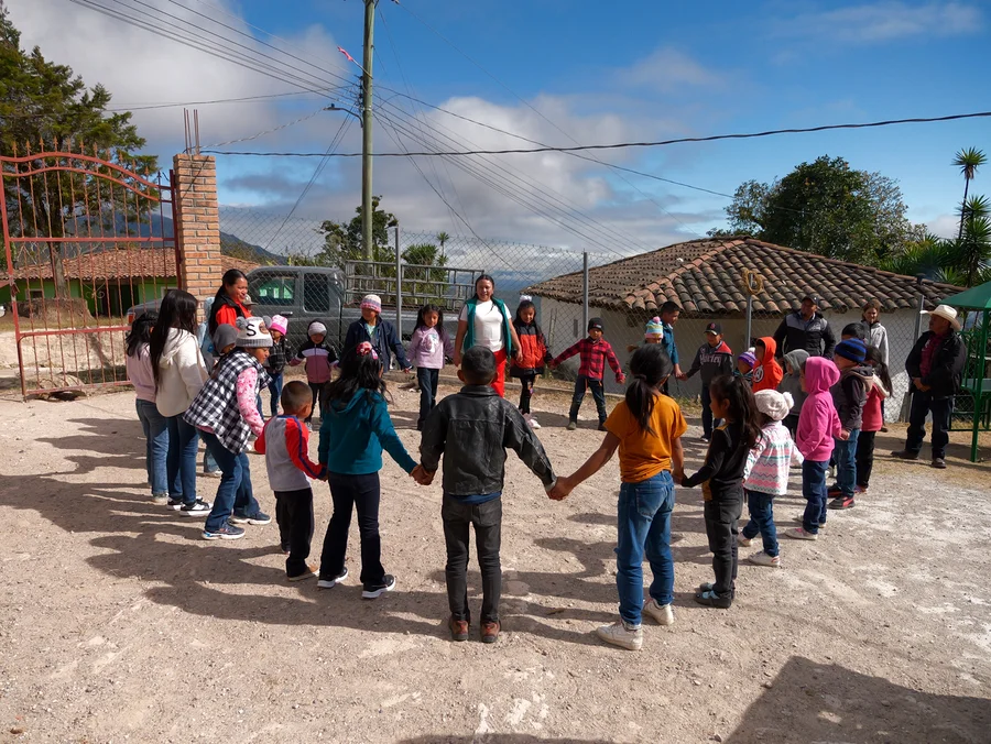 Eine Gruppe Kinder und Erwachsene hält sich im Kreis auf einem Feldweg im Freien an den Händen. Im Hintergrund sind ein rotes Tor, ein Lastwagen und ein Gebäude mit Ziegeldach zu sehen.