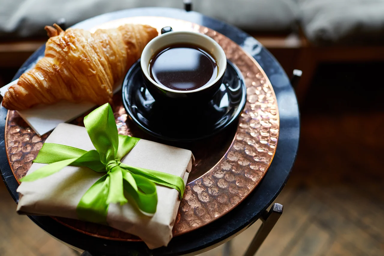 Auf einem Tisch liegen ein Croissant, eine Tasse Kaffee und ein verpacktes Geschenk mit grünem Band.