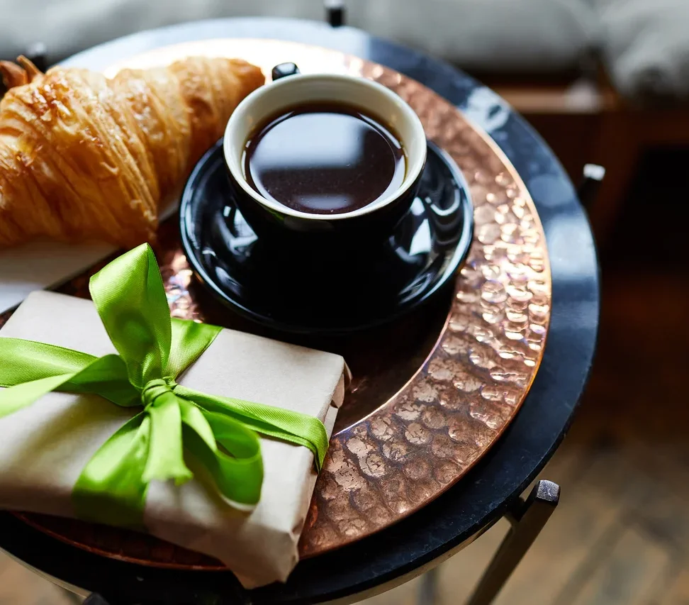 Auf einem Tisch liegen ein Croissant, eine Tasse Kaffee und ein verpacktes Geschenk mit grünem Band.