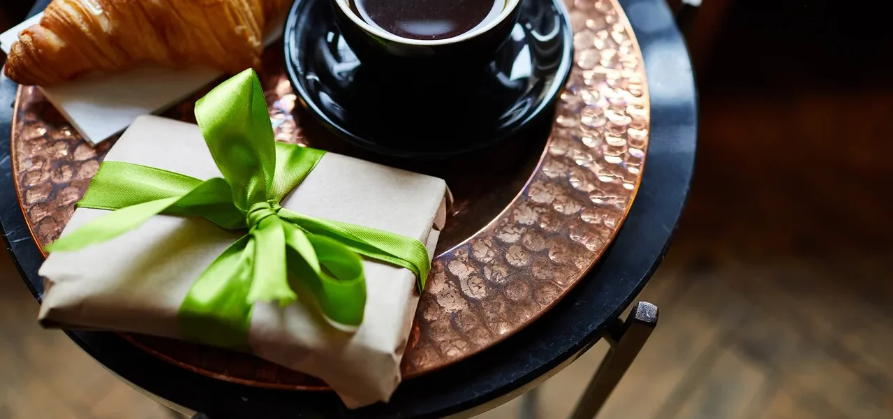 Auf einem Tisch liegen ein Croissant, eine Tasse Kaffee und ein verpacktes Geschenk mit grünem Band.