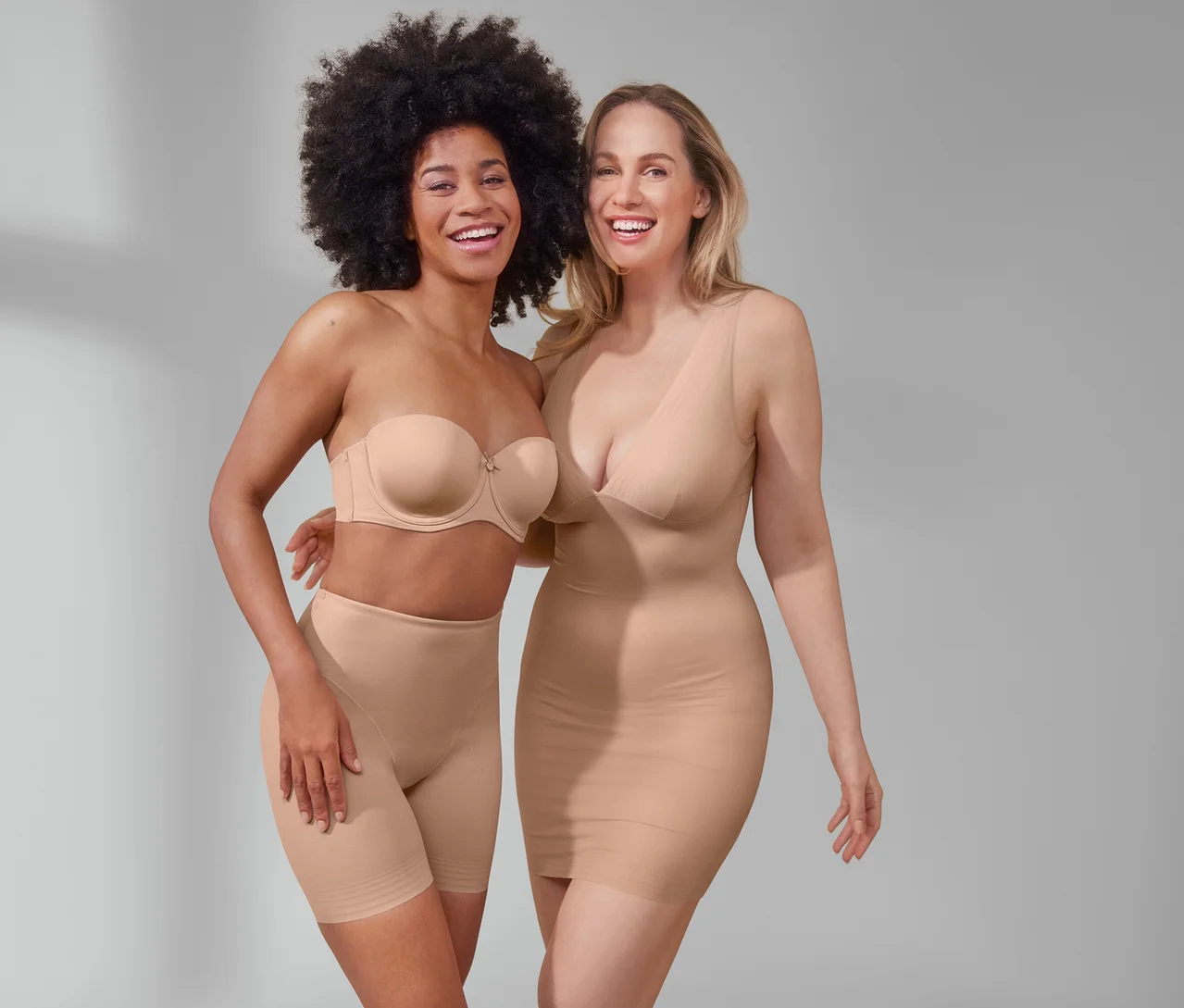 Zwei lächelnde Frauen posieren. Eine trägt einen Multistyle-BH und beige Shorts »Shaping«. Die andere trägt ein beiges Unterkleid »Shaping«.