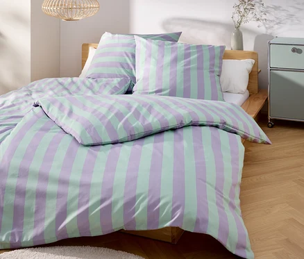 Ein Bett mit gestreifter Bettwäsche in Lila und Grün steht in einem hellen Raum.